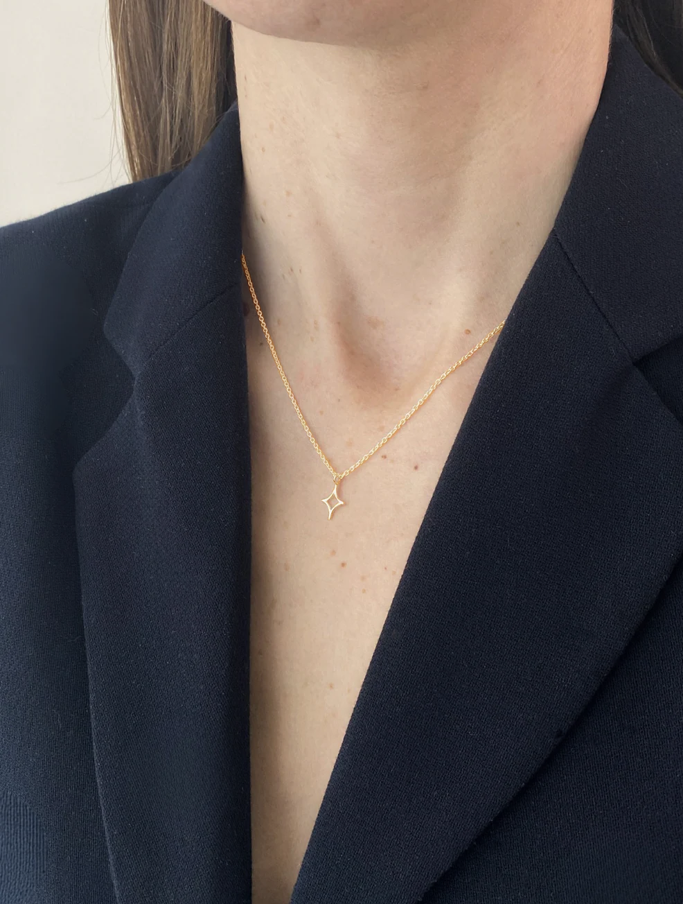 Person wearing a delicate necklace with a small pendant against a dark blazer.